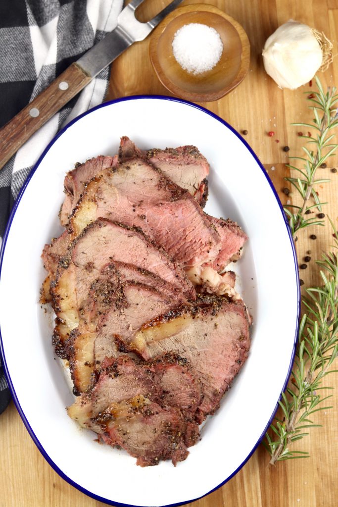 Sliced roast beef on a blue rimmed white platter, meat fork, bowl of salt, whole garlic clove, peppercorns and fresh rosemary.