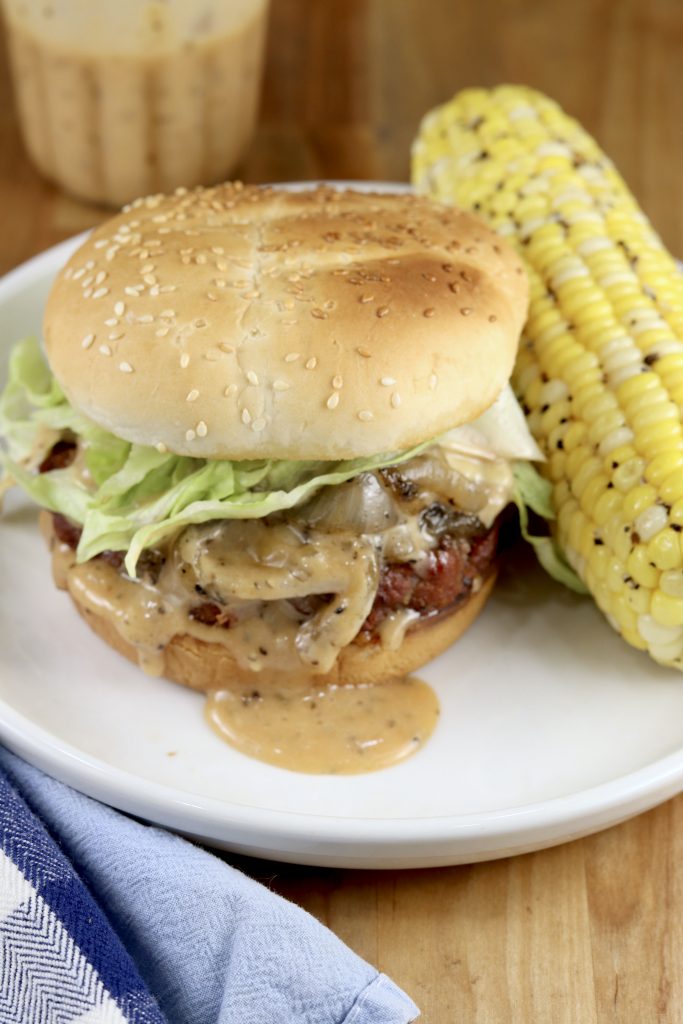 Grilled Onion Burger with burger sauce, toasted bun and a side of grilled corn on the cob on a white plate 