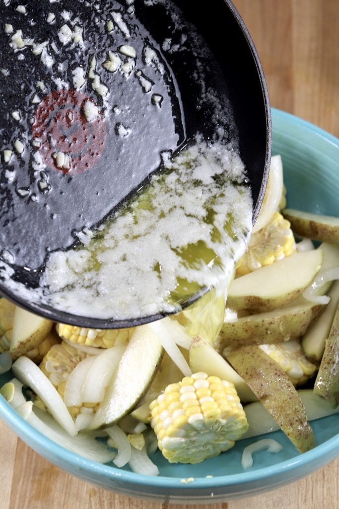 pouring melted butter into a bowl of corn cobs, potatoes and onions