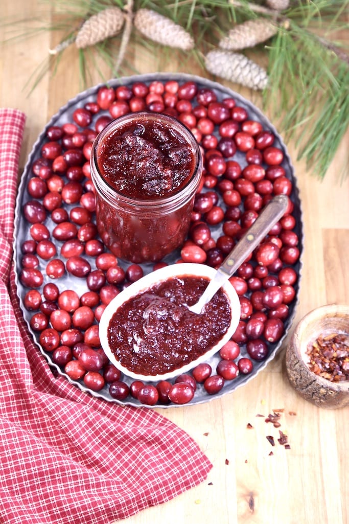 Overhead view of tray of fresh cranberries with jar of Cranberry BBQ Sauce and in a bowl with a spoon. Pine cones and boughs in background