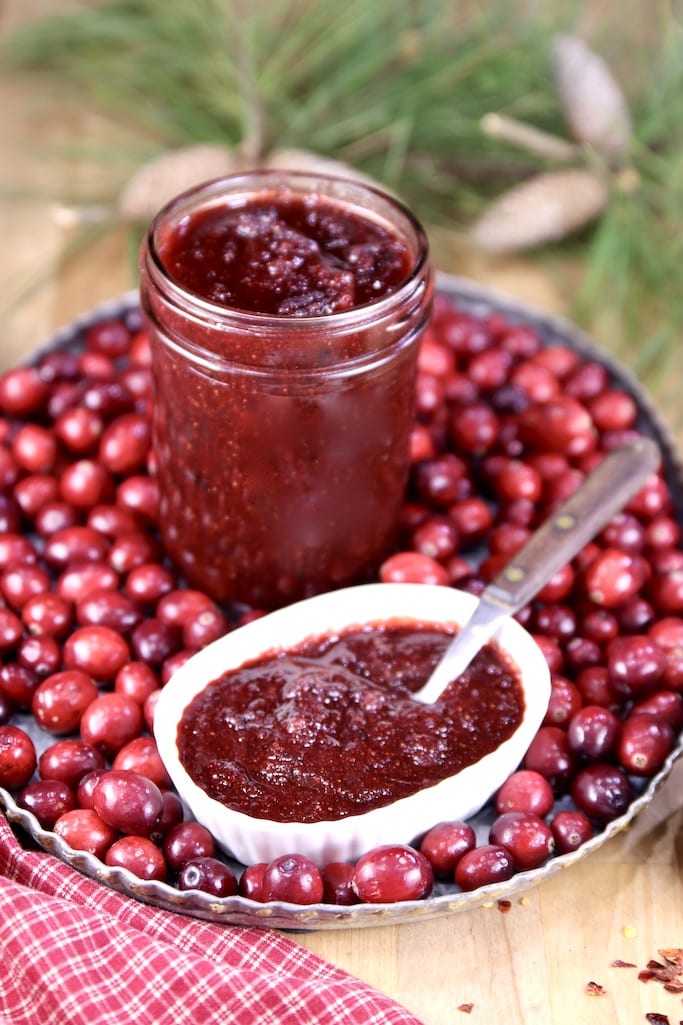 Cranberry BBQ Sauce in a jar and small bowl with a spoon. All on a tray of fresh cranberries. Pine boughs in background