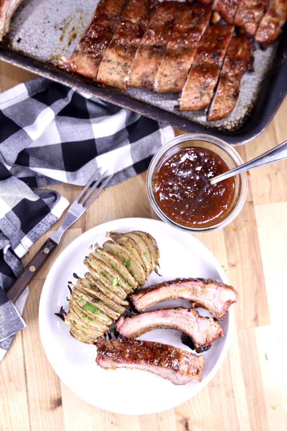 plate with 3 baby back ribs with a potato, jar of bbq sauce and pan of ribs in background