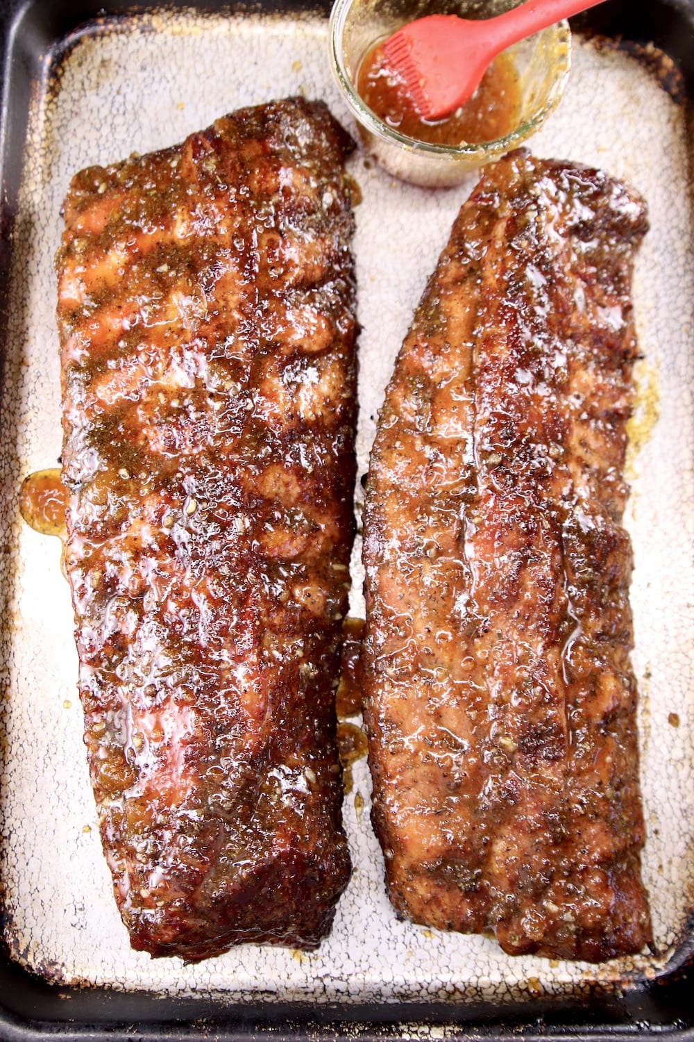2 racks of ribs brushed with bbq sauce on a sheet pan with jar of sauce and brush