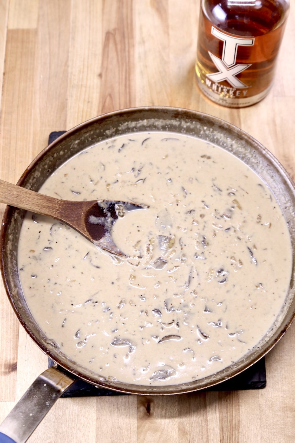 pan of creamy mushroom sauce with a wood spoon, bottle of whiskey to the side