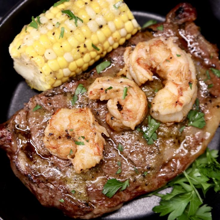 Grilled Ribeye Steak topped with 3 grilled shrimp, served with ½ corn on the cob and parsley garnish on a black plate