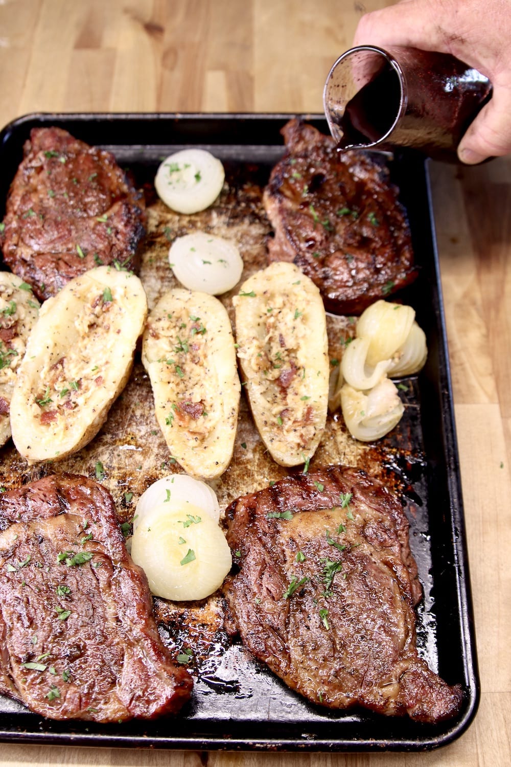 drizzling steak sauce over grilled ribeyes on a sheet pan with baked potato halves
