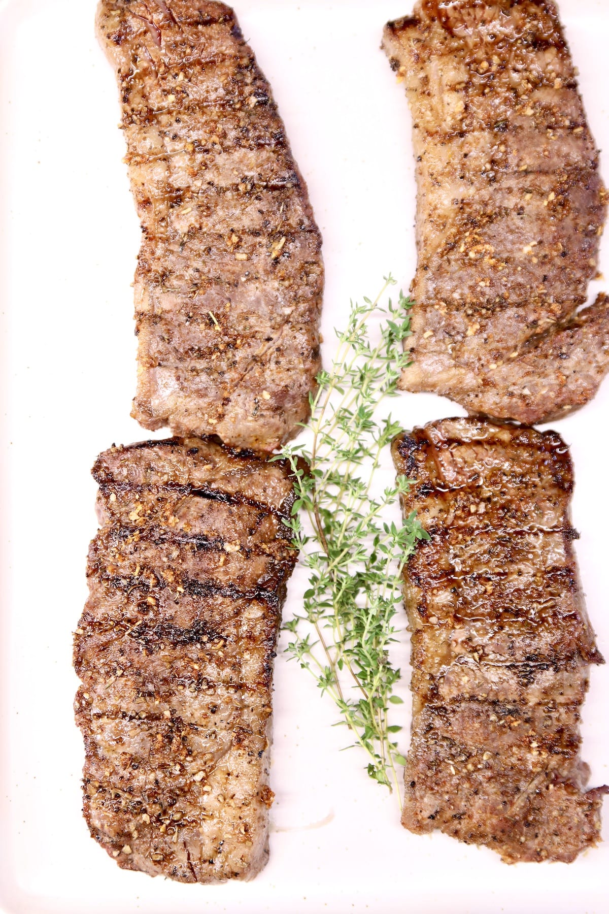 4 grilled strip steaks on a white tray with fresh thyme in center