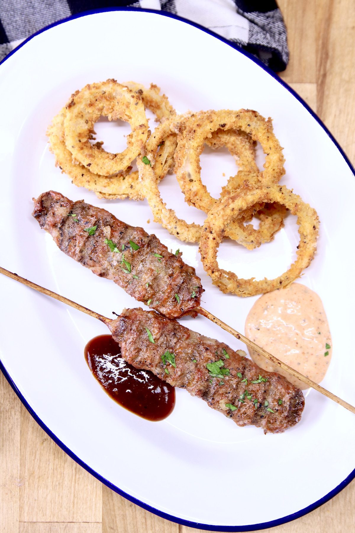 2 beef kabobs on an oval platter with onion rings and 2 dipping sauces