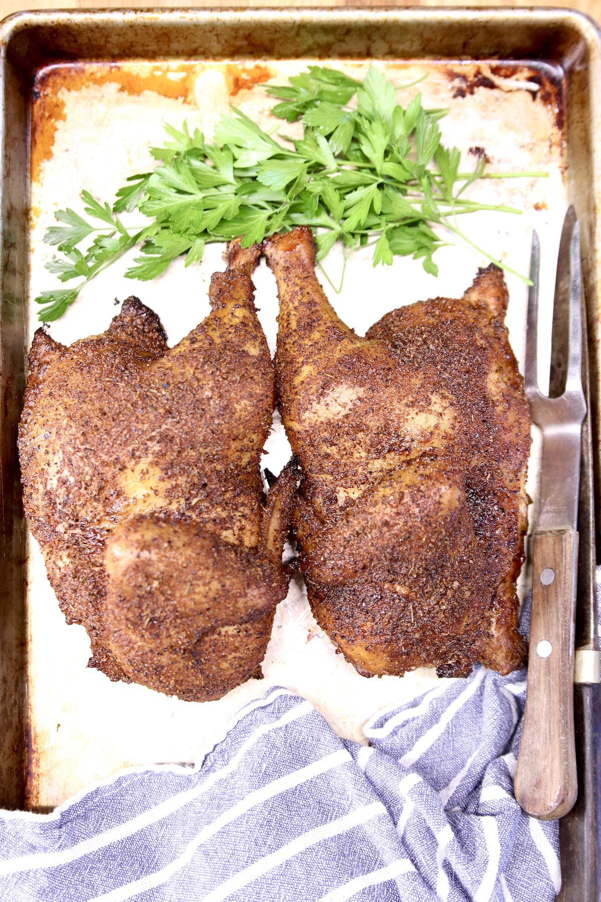 Grilled half chicken on a sheet pan with parsley, blue towel, meat fork and knife