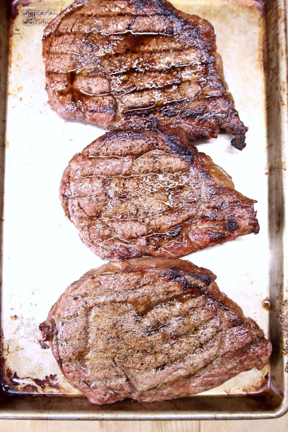 3 grilled steaks on a rimmed baking sheet.