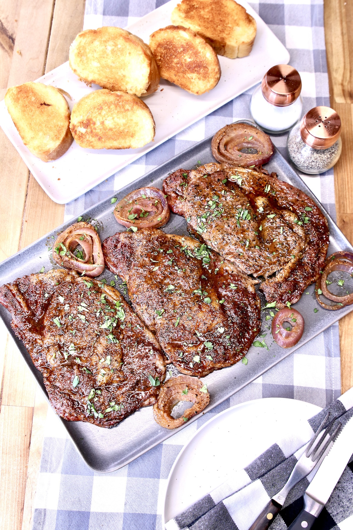 Grilled ribeyes on a platter, garlic bread in the background.