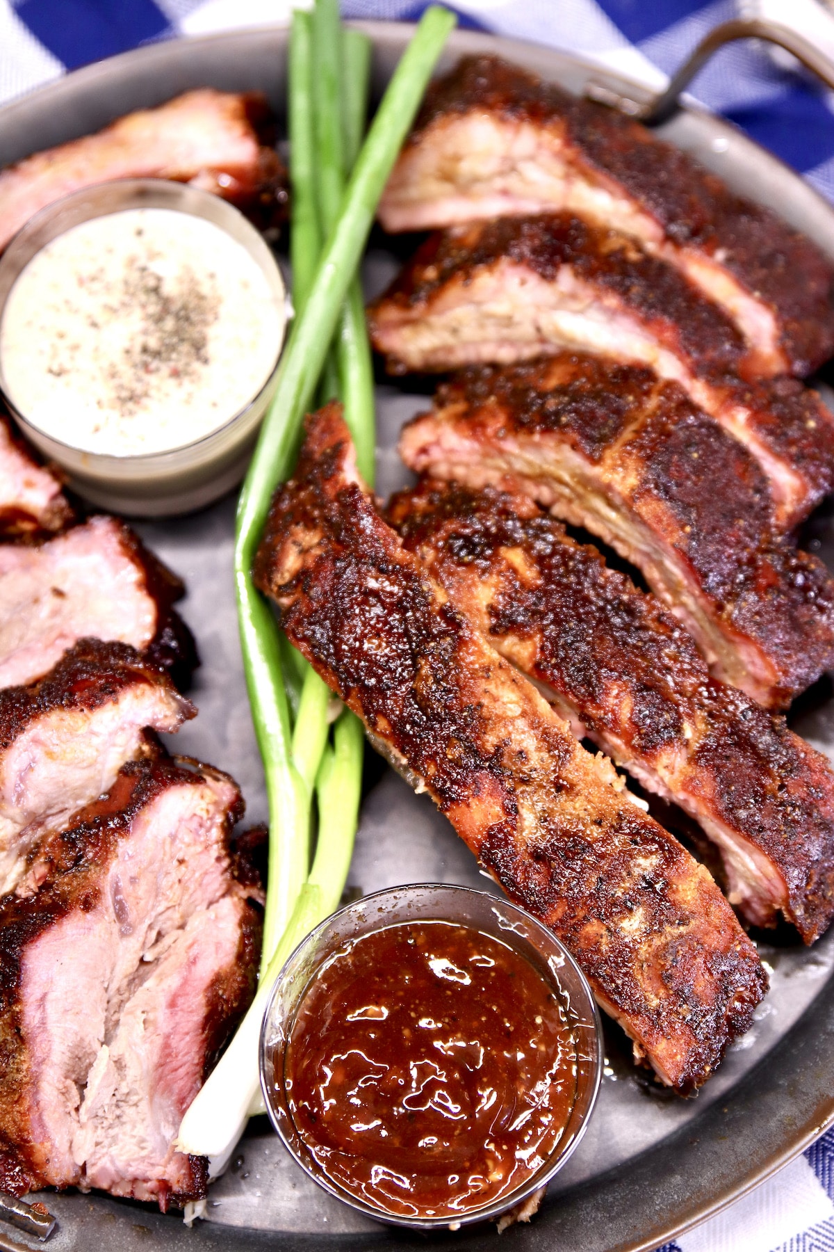 Closeup of platter of sliced baby back ribs, green onions, bowls of sauce.