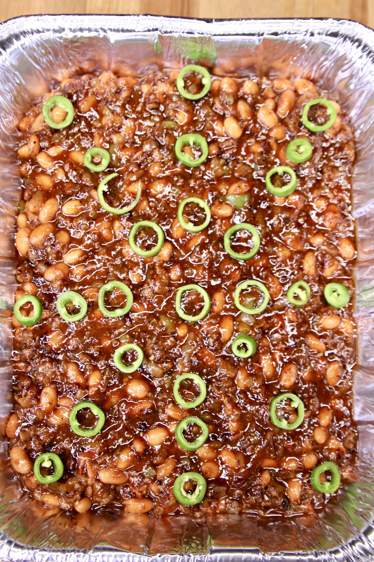 Cowboy beans with jalapeno slices in a foil pan.