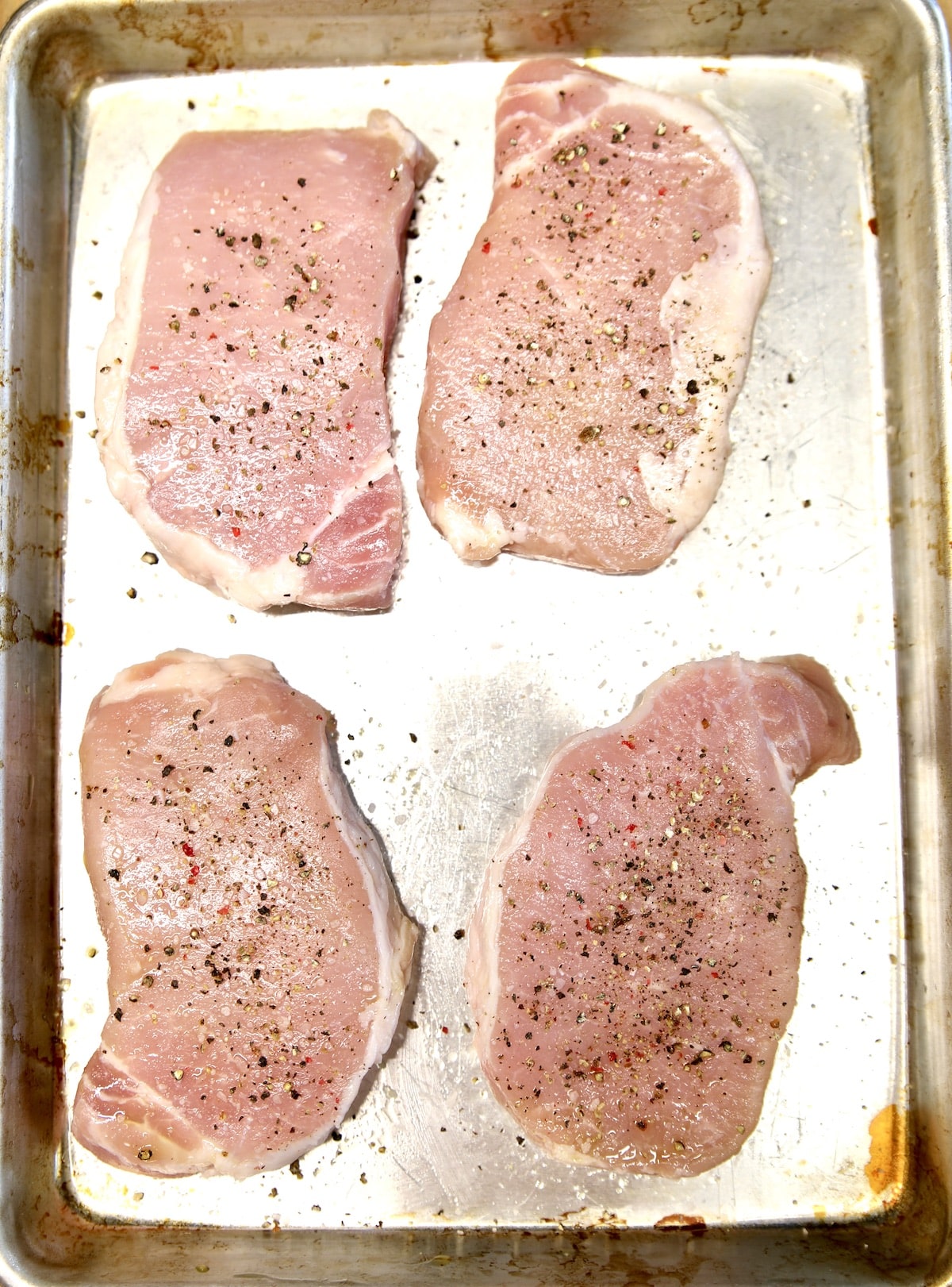 4 raw pork chops on a baking sheet, seasoned.