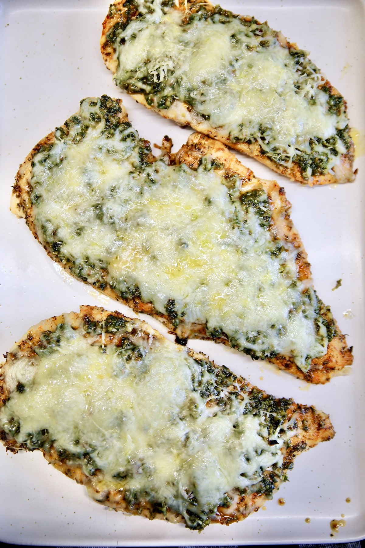3 chicken cutlets with pesto and melted cheese on a pan.