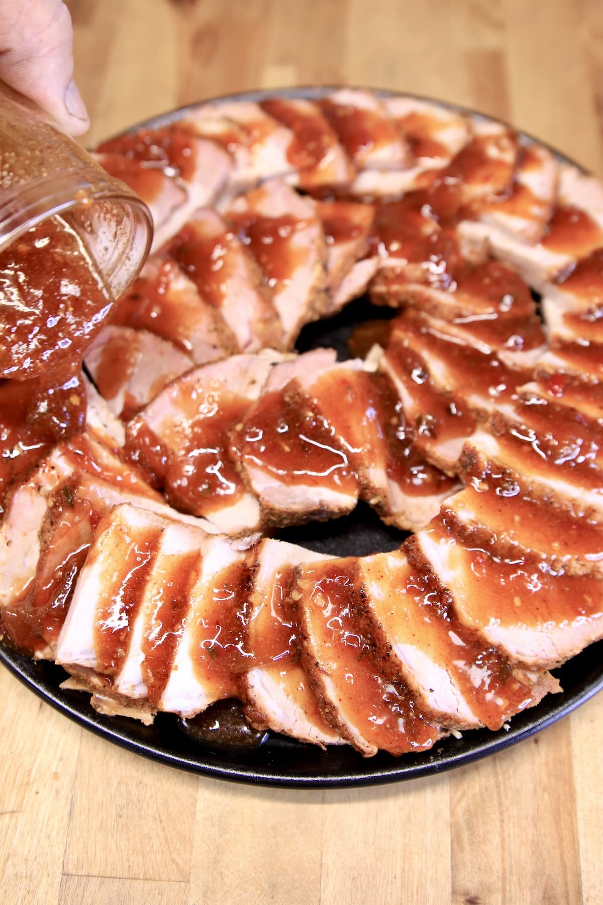 Pouring bbq sauce over sliced pork loin on a round platter.