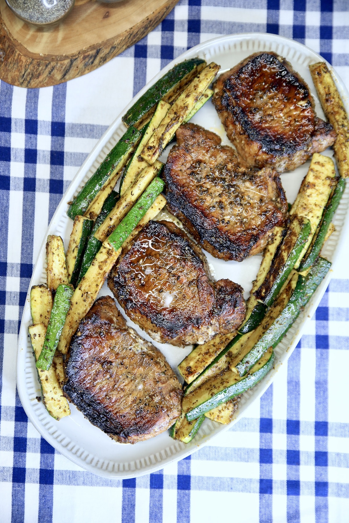 Grilled pork chops on a platter with zucchini on a blue and white plaid cloth.