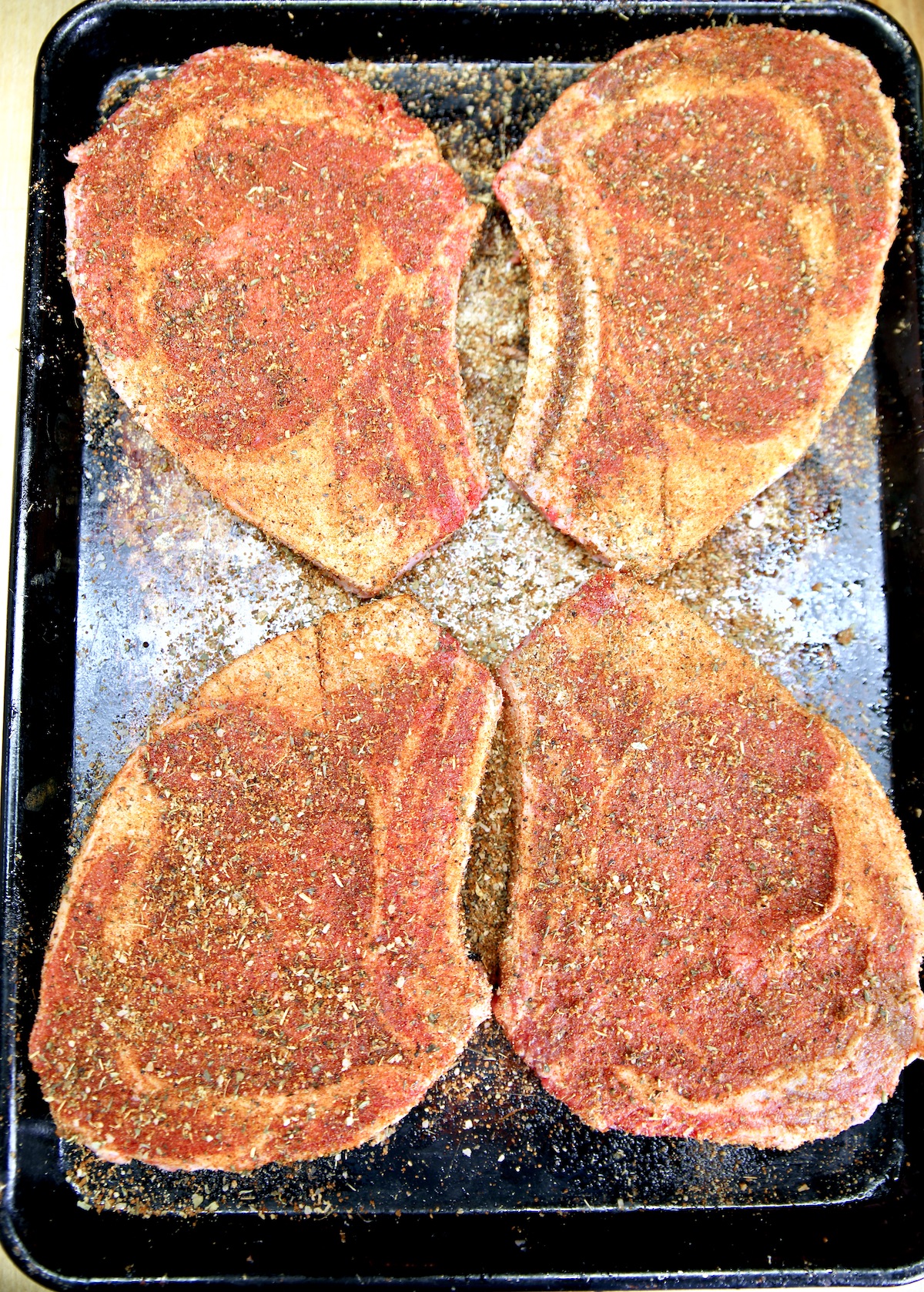 4 bone-in ribeye steaks on a sheet pan seasoned with dry rub.