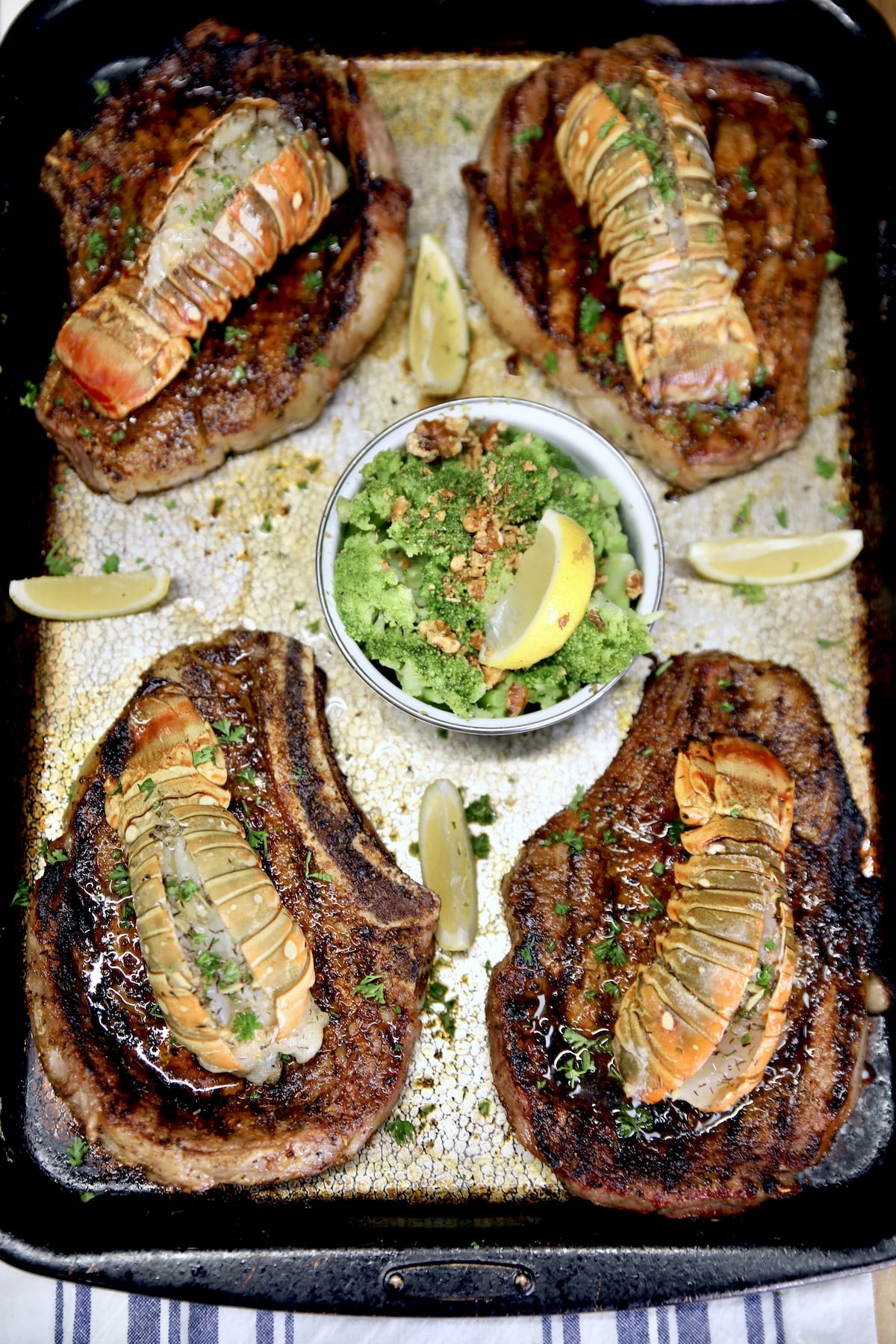 Pan of 4 steaks with lobster tails. Bowl of broccoli, lemon slices.