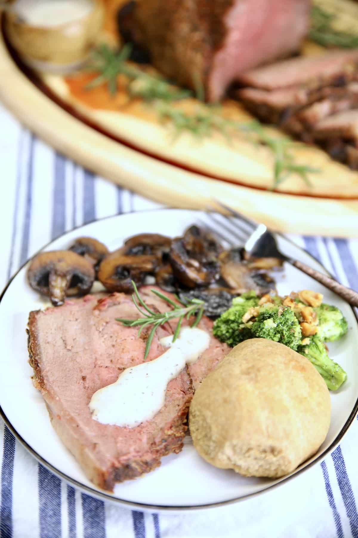 Plate with slice of roast beef with horseradish sauce, mushrooms, broccoli & dinner roll.