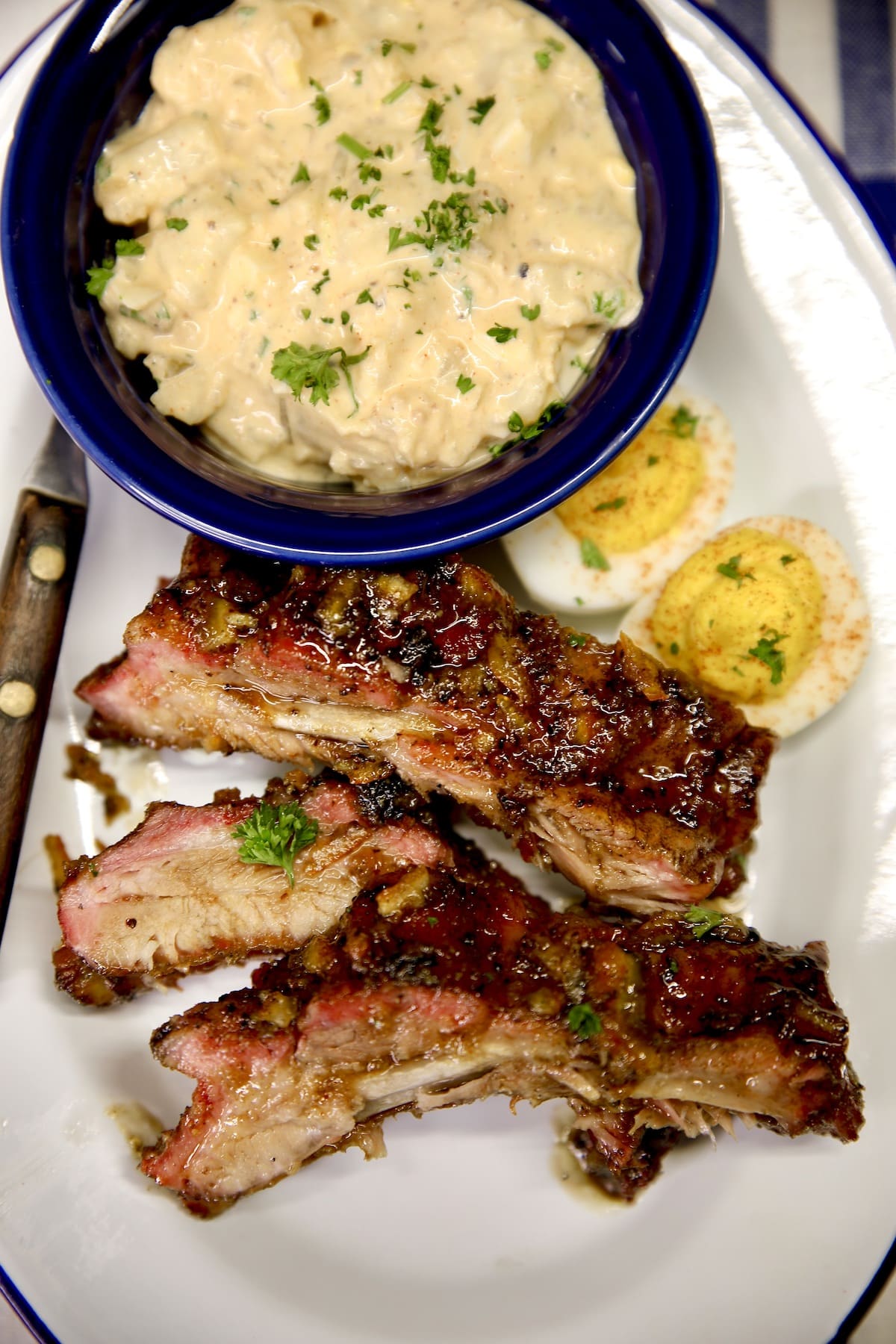 3 ribs on a plate with bowl of potato salad and deviled eggs.