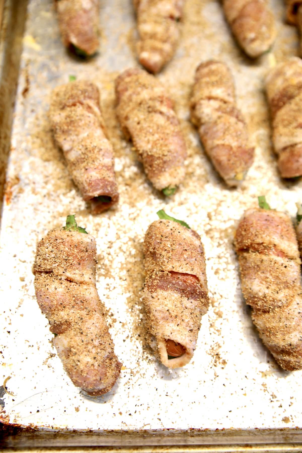 Uncooked jalapeño poppers wrapped in bacon with dry rub seasoning.