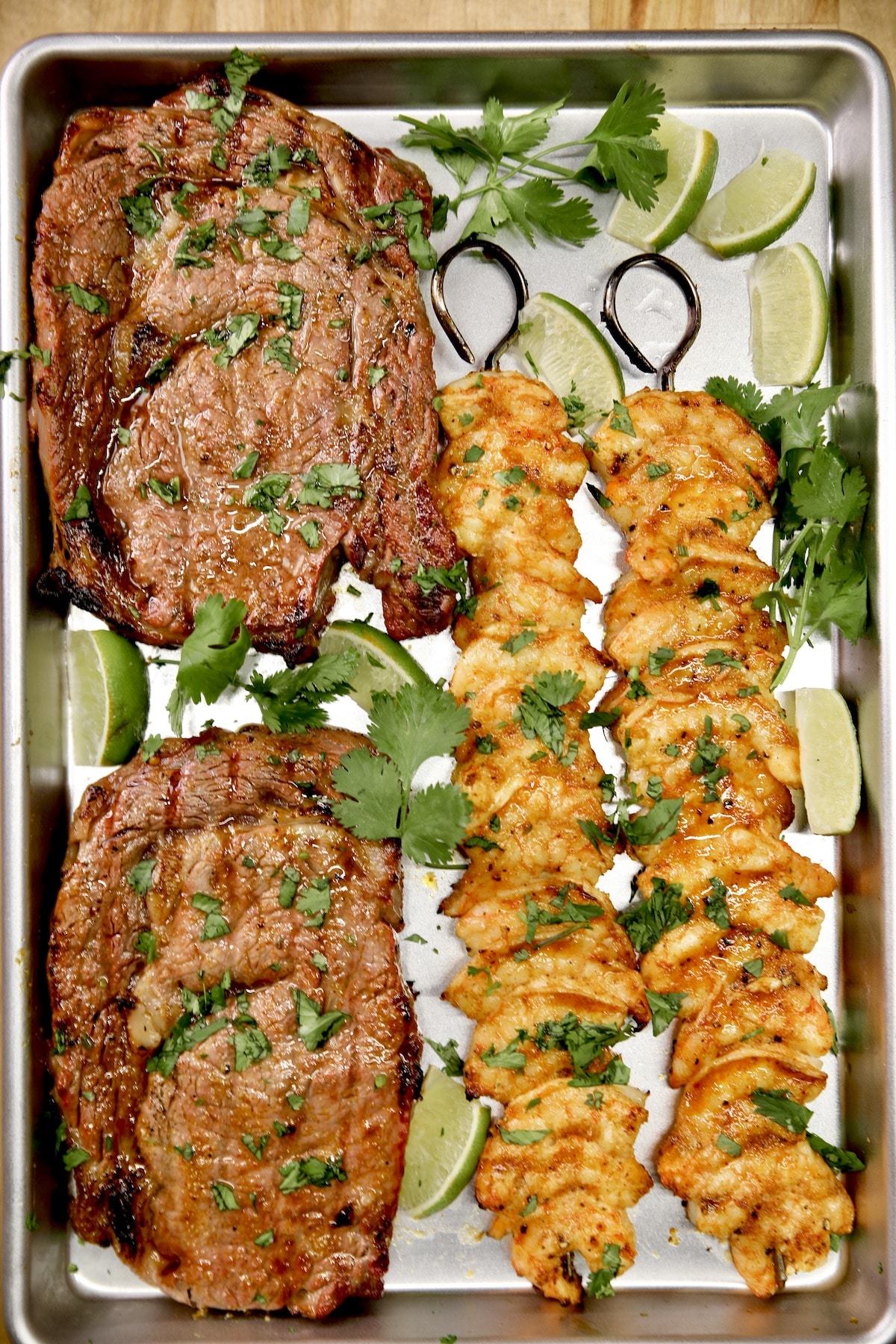 Grilled steaks and shrimp skewers with cilantro and lime on sheet pan.