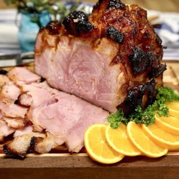 Orange Glazed ham, partially sliced on a cutting board with orange slices & parsley.