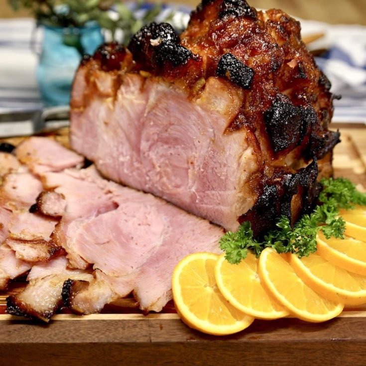 Orange Glazed ham, partially sliced on a cutting board with orange slices & parsley.