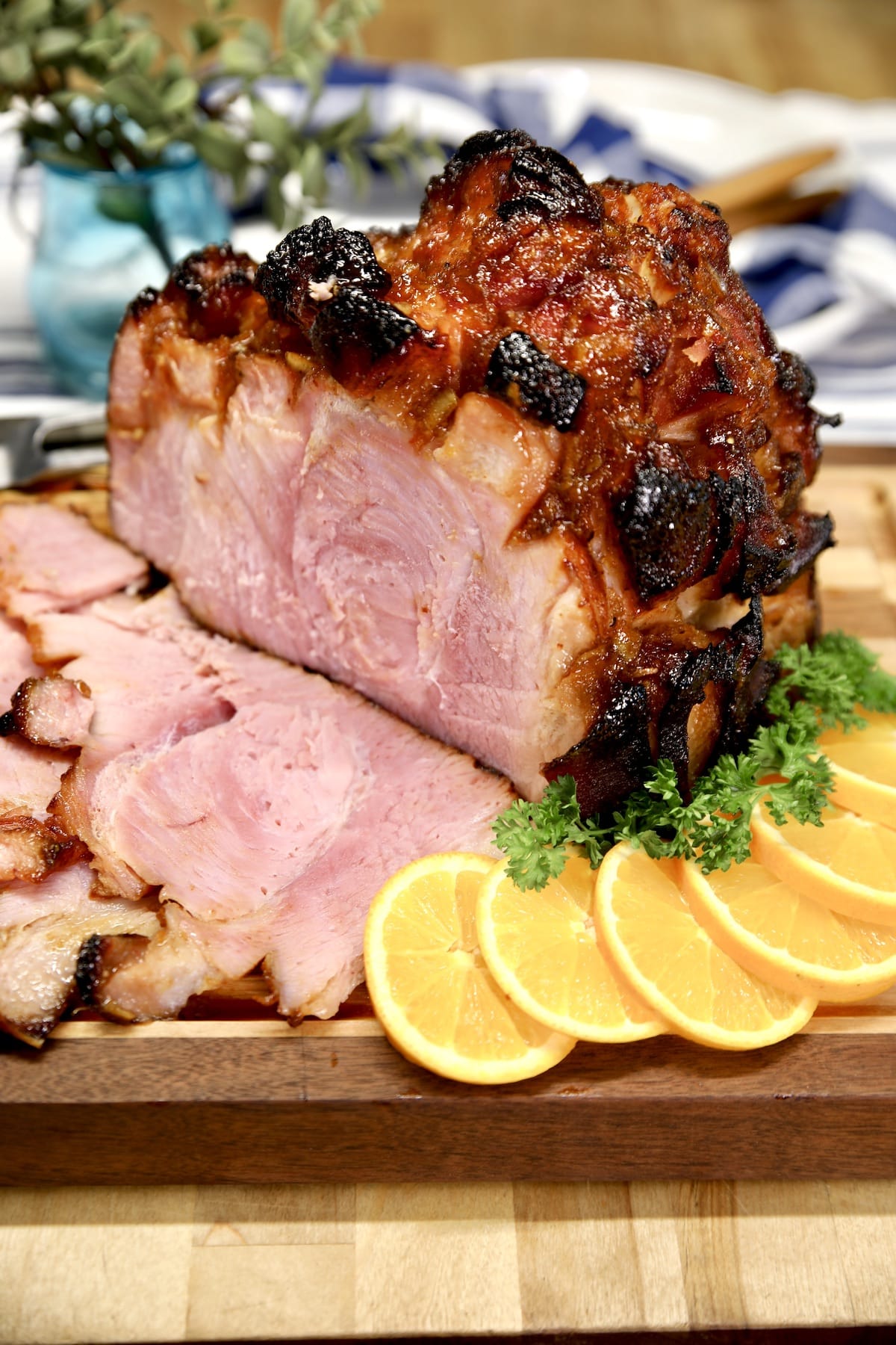 Orange glazed ham, partially sliced on a cutting board with orange slices and parsley.