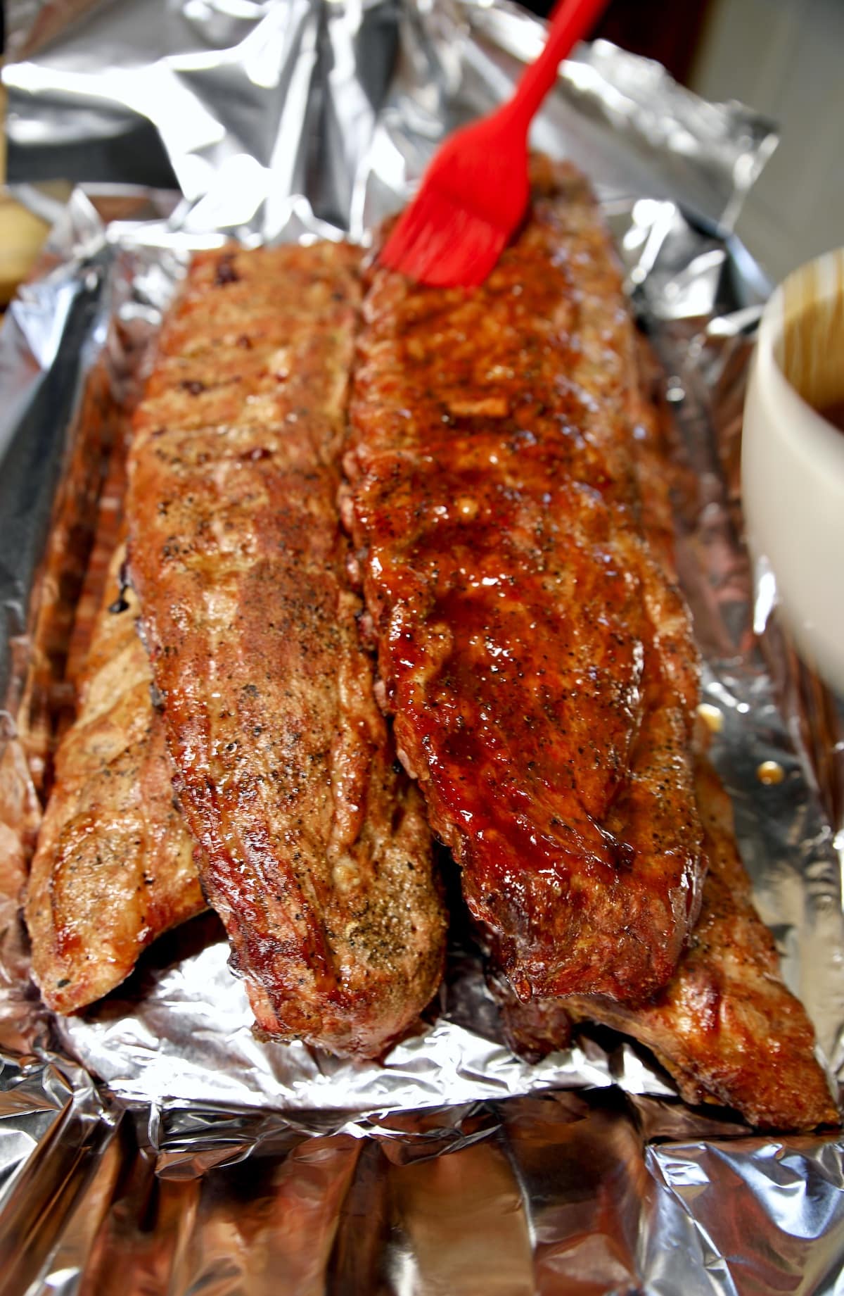 Smoked baby back ribs on a foil lined pan, brushing with sauce.