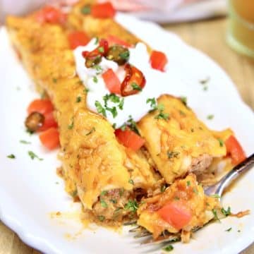 Pulled pork enchiladas on a plate with sour cream and tomatoes.