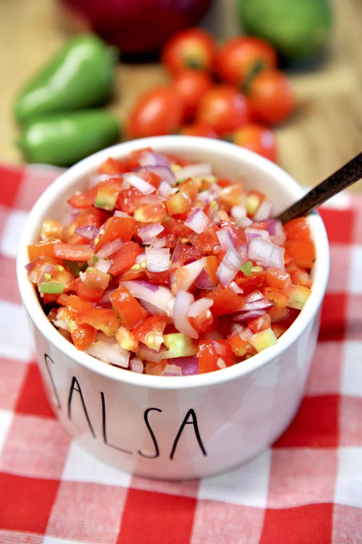 Bowl of fresh salsa with a spoon, vegetables in the background.