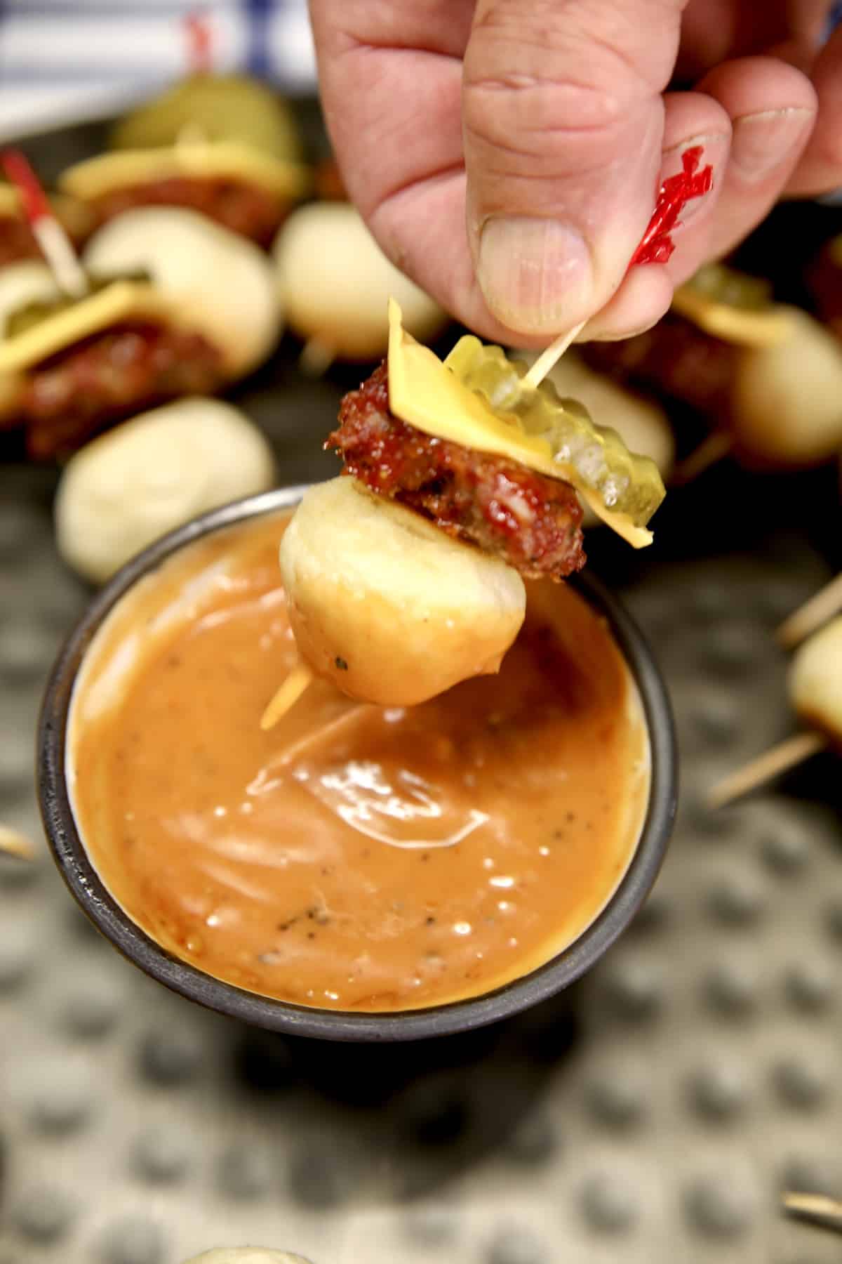 Mini cheeseburger appetizers on a stick, dipping into sauce.