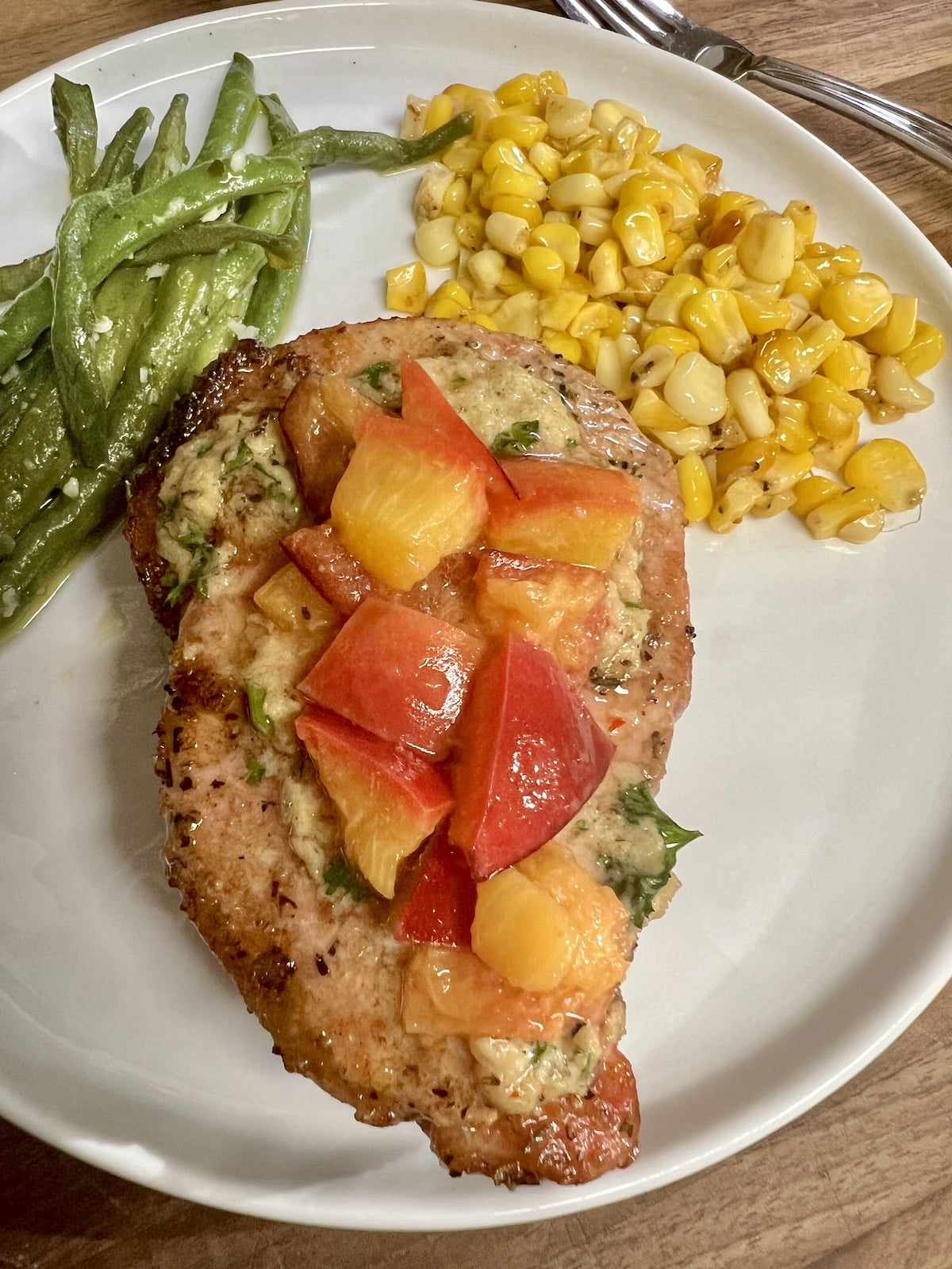 Pork chop with grilled peaches on a plate with corn and green beans.