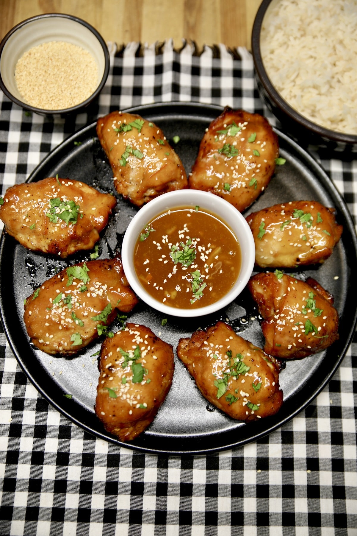 Platter of chicken thighs with bowl of orange sauce, rice and sesame seeds.