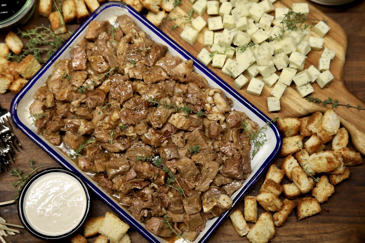 Charcuterie board with steak bites in a pan, horseradish sauce, cheese bites and bread cubes.