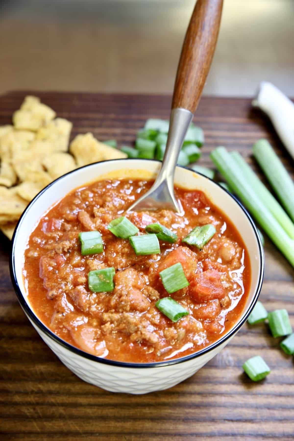Chili in a bowl with a spoon on a wood board with fritos and green onions.