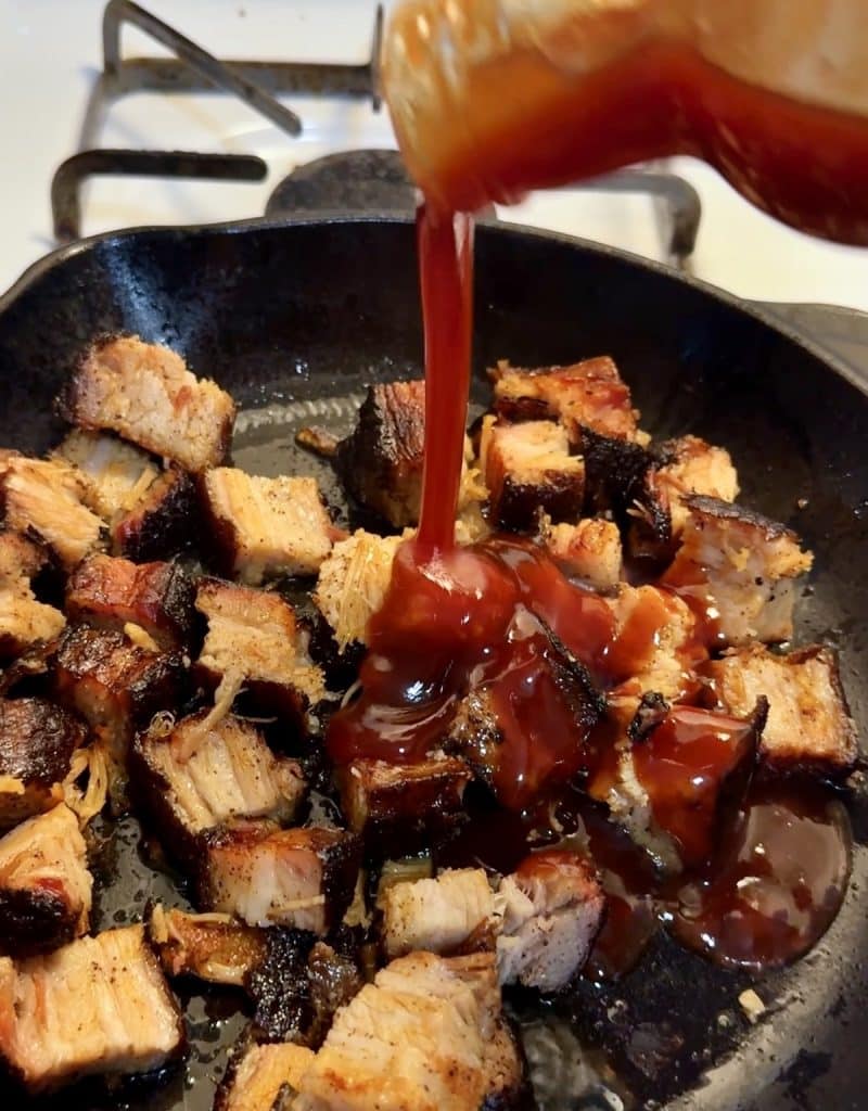 Pork belly burnt ends in a skillet with bbq sauce pouring over.